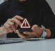 A imagem mostra um homem segurando um smartphone e em frente a um notebook. Acima há o símbolo de alerta/perigo. 
